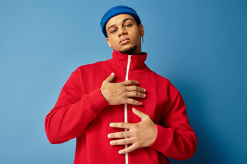 Trendy young man in a beanie and red jacket poses against a blue backdrop