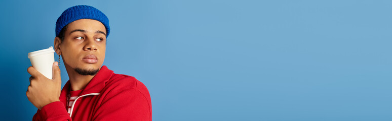 Stylish young man in trendy beanie and red jacket poses against a blue backdrop