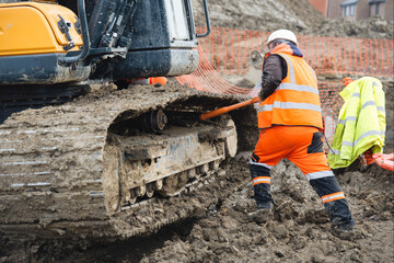 Construction worker  in bright safety gear working hard using shovel to clean tracks of his heavy machinery excavator on muddy site © Iryna
