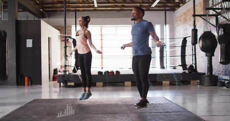 Jumping fitness duo synchronizing rope jumps in fitness studio, with punching bags and kettlebells