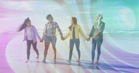 Walking four friends in jackets holding hands along beach at low tide, with sunlight lens flare