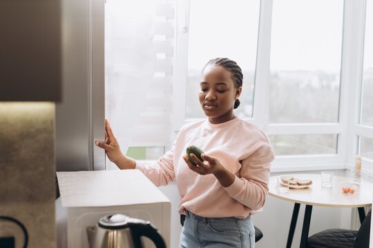 Woman holding avocado and opening refrigerator in kitchen - Powered by Adobe