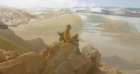 Perching hiker wearing outdoor gear on cliff overlooking tidal flat, with backpack and sleeping mat