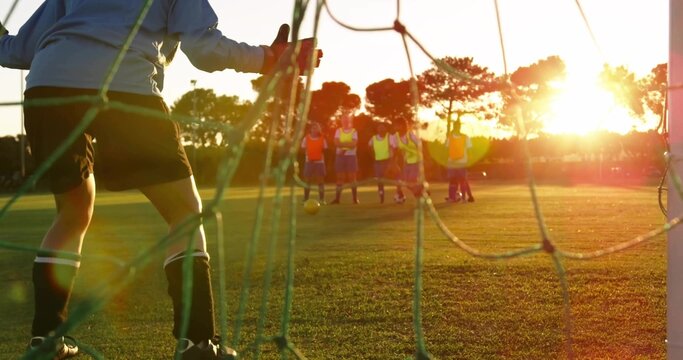 Steadying goalkeeper bracing on goal net at dusk training field, with ball, gloves, bibs and cleats