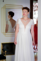 Fototapeta premium A woman in a white dress is standing. There is a mirror behind the woman. The woman's reflection in the mirror is visible.