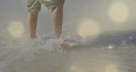 Standing adult male wearing rolled-up shorts in shallow ocean water, sunlight creating flares