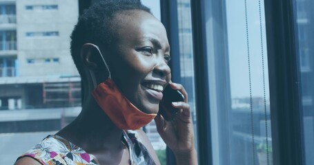 Employee wearing floral blouse in office near window blinds, talking on smartphone with mask down