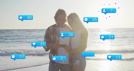Couple checking smartphone notifications on beach at sunset, with social media icons
