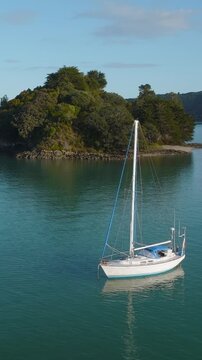 A sailboat is anchored in calm, turquoise water near a small, tree-covered island in New Zealand. The boat is white with blue trim and a tall mast. Coromandel Peninsula, Oamaru Bay