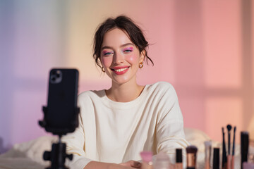 Smiling beauty influencer recording a makeup tutorial with smartphone in a pastel studio, surrounded by brushes and cosmetics