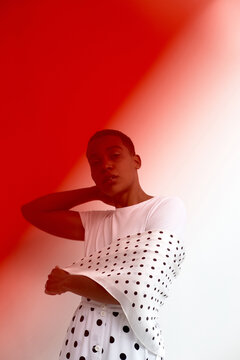 Portrait of young Black woman standing with hand behind neck, wearing polka dot garment, looking into camera against gradient red and background, modern fashion concept