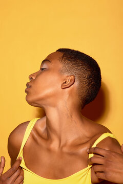 Young Black woman turning head to side, exposing neck and jawline, holding strap of top with hand, short hair visible, posing against plain yellow background