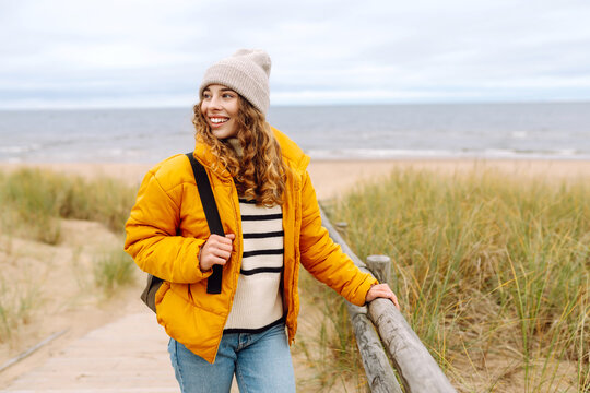 A happy woman in a bright yellow jacket stands on a sandy beach. The traveler enjoys the seascape and walks at sunset. Travel, recreation concept. Active lifestyle.