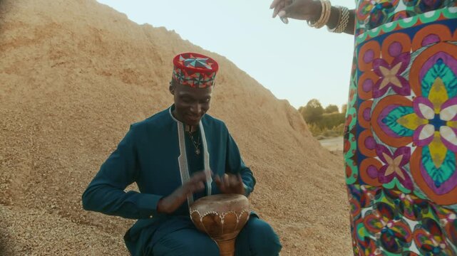 Young Black man wearing ethnic outfit with kofia sitting by fire, playing goblet drum for dancing woman