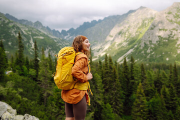 A female hiker with a yellow backpack on a rocky path on a sunny day. Happy woman enjoying the mountain landscape and feeling freedom in the open air. Nature, adventure concept.