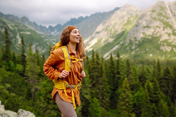 Naklejka premium A female hiker with a yellow backpack on a rocky path on a sunny day. Happy woman enjoying the mountain landscape and feeling freedom in the open air. Nature, adventure concept.