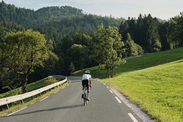 Fototapeta premium A cyclist rides along a scenic country road, surrounded by lush green fields and forests. The image evokes a sense of freedom and tranquility, ideal for travel, sports, or nature-themed projects.