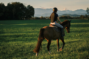 Woman horseback riding in a lush green field with mountains in the background.  Golden hour light...