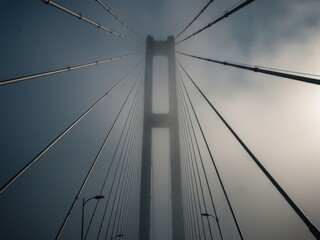 A bridge tower emerges from dense fog, framed by its converging cables, creating a dramatic, ethereal, and architectural scene against a hazy sky.