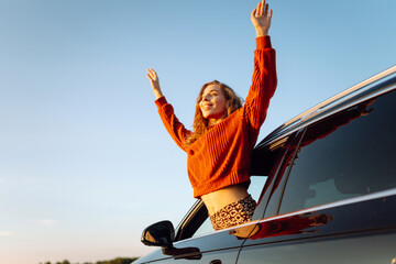 Beautiful woman leaning out of car window catching wind in her hair. Young woman feeling freedom from car window enjoying sun rays on journey. Car travel concept, adventure and freedom. © maxbelchenko