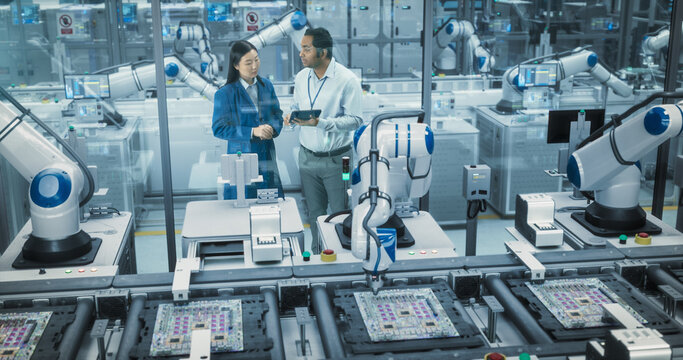 Two Diverse Engineers Talking on a Factory Floor, Using Tablet Computer at an Electronics Manufacturing Plant. Female and Male Specialists Chatting, Monitoring Robots on a Modern Assembly Line