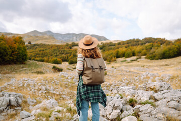 Fototapeta premium A rear view of a female hiker with a backpack and a hat walking along a rocky trail at sunset. Young woman enjoying the mountain scenery, admiring the views. Adventure, freedom and weekend concept.