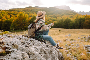 Fototapeta premium A rear view of a female hiker with a backpack and a hat walking along a rocky trail at sunset. Young woman enjoying the mountain scenery, admiring the views. Adventure, freedom and weekend concept.