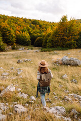 A rear view of a female hiker with a backpack and a hat walking along a rocky trail at sunset. Young woman enjoying the mountain scenery, admiring the views. Adventure, freedom and weekend concept.