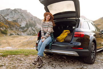 Naklejka premium A joyful woman in a hat sits in the trunk of a car and admires the mountain landscape. A woman on a car trip enjoys the sunset and mountains. Transport concept, adventure.