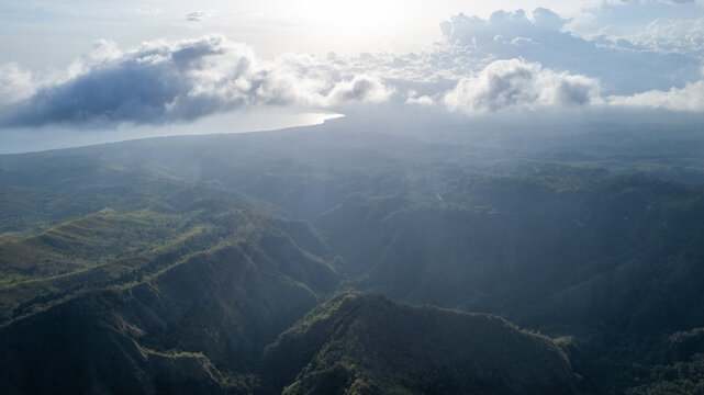 Drone aerial view of dramatic mountains and valleys with clouds drifting above and the ocean horizon glowing under the sunlight, showcasing the harmony of land, sea, and sky.