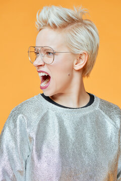 Fashion model with emotional grimace. Profile portrait of young stylish Caucasian woman with spiked up blond hair  grimacing with open mouth on orange background