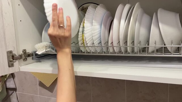 Hand reaches in to take out a plate. Close-up view of kitchen cabinet opening to reveal dish drying rack filled with various plates. Cabinet doors are light-colored with wood grain texture.