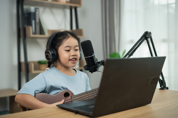 Fototapeta premium Young girl with headphones sings and plays ukulele in front of laptop during online music lesson in modern home setup with natural light.