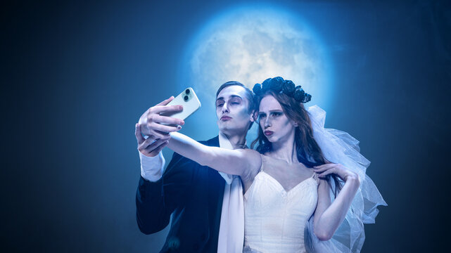 Gothic couple taking eerie selfie with smartphone under full moon. Concept of surreal gothic humor, Halloween fashion, dark romance, horror cinema, and bizarre editorial fantasy.