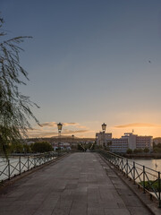 Pedestrian bridge over the Tua River in Mirandela, Portugal, leads to the city center at sunset. Evening light, calm water, and urban skyline create a tranquil and scenic connection.