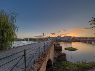 Pedestrian bridge over the Tua River in Mirandela, Portugal, leads to the city center at sunset. Evening light, calm water, and urban skyline create a tranquil and scenic connection.