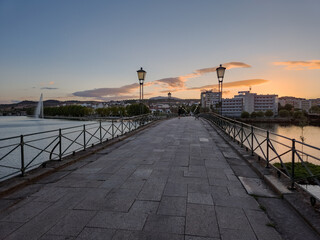 Pedestrian bridge over the Tua River in Mirandela, Portugal, leads to the city center at sunset. Evening light, calm water, and urban skyline create a tranquil and scenic connection.