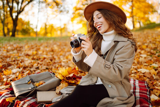 Woman is sitting on blanket in park covered with autumn leaves with camera in hands. Beautiful woman in hat is enjoying autumn landscape and taking pictures on camera. Concept of beauty, relaxation.