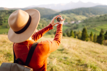 Rear view of female hiker with backpack and hat holding compass against hills on sunny day. Female hiker looking for hiking direction, enjoying sunset mountain landscape. Navigation concept.