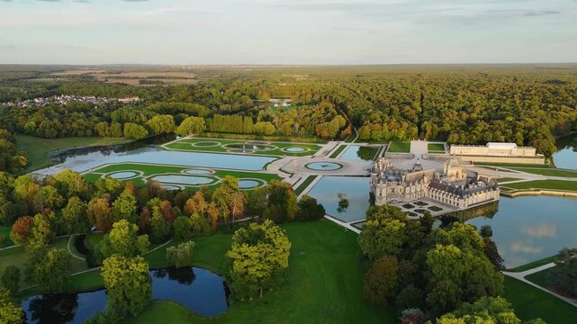 Aerial drone view captures Ch&acirc;teau de Chantilly in France during golden hour sunset, with slow circular motion over formal gardens and Grand Bassin lake
