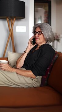 Senior woman relaxing on sofa, talking on phone and drinking tea