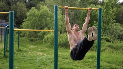 Fototapeta premium Athletic man trains abdominal muscles raising legs on horizontal bar at sports ground. Young sportsman exercising on pull-up bar outdoor. Sporty athlete working out at nature. Healthy lifestyle