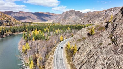 A panoramic aerial view of a car driving on a winding road alongside a wide river, with surrounding mountains covered in vibrant golden autumn foliage under a clear blue sky - Powered by Adobe