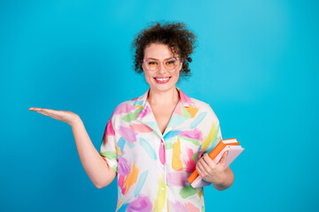 Cheerful woman wearing a colorful shirt gestures with one hand while holding books against a blue background