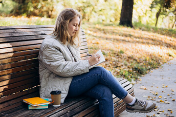Young woman writing in notebook on bench in autumn park.