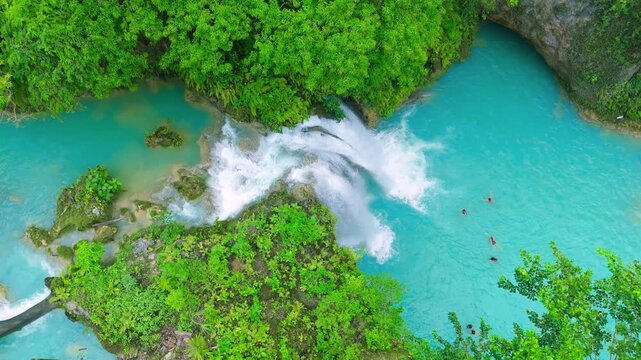 Aerial view of multi-tiered waterfalls cascading into vibrant turquoise pools, surrounded by dense green jungle foliage with people swimming, Kawasan Falls, Philippines.