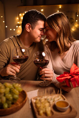Happy young couple in love hugging holding glasses having romantic dinner date with candles in dark light, drinking wine, celebrating Valentines day at home.