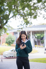 Fototapeta premium Asian female student in warm clothes using phone to chat with friends and ready for video call while in university.