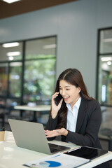 Fototapeta premium A beautiful businesswoman in a suit is looking at a computer screen while talking on her mobile phone. She is sitting in an office and using modern technology.