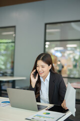 Fototapeta premium Asian businesswoman working at desk using laptop and notebook, talking with client or modern office building.